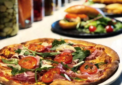 Pizza topped with cherry tomatoes, arugula, ham, and shaved parmesan on a golden crust.